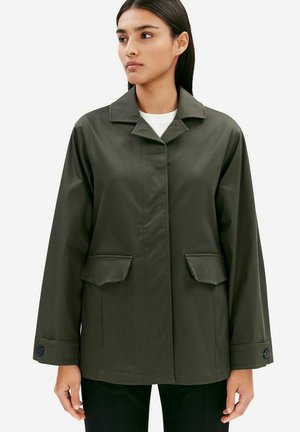 Woman wearing an olive green jacket with buttoned cuffs and two flap pockets, standing against a plain white background.