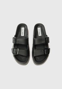 Juodos šlepetės su dviem reguliuojamais dirželiais su sagtimis, tekstūruotu padu ir lygiu išoriniu paviršiumi. Viduje pažymėtos "Steve Madden".