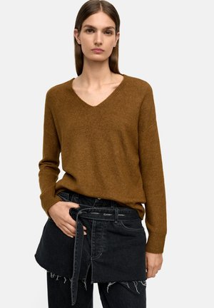 Brązowy sweter w serek z długimi rękawami, wykonany z miękkiego materiału. Połączony z czarnymi dżinsami o warstwowym kroju i postrzępionym wykończeniu.