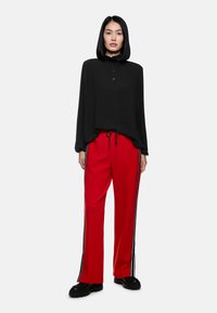 Schwarzes Langarmshirt mit Knöpfen, kombiniert mit roten weit geschnittenen Hosen mit schwarzen Seitenstreifen. Schwarze klobige Schuhe vervollständigen das Outfit.