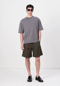 T-shirt gris surdimensionné à manches courtes, shorts verts olive, mocassins noirs et lunettes de soleil. Tenue décontractée avec une coupe ample et des couleurs unies.