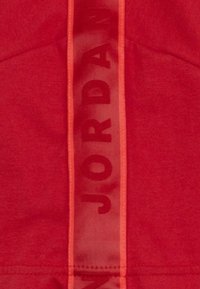 Tessuto atletico rosso con una sovrapposizione semi-trasparente caratterizzata dalla parola "JORDAN" in verticale di un rosso più scuro, dettagli ricamati e una texture liscia.