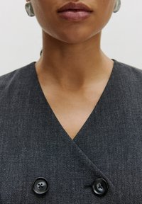 Graues, doppelt geknöpftes Kleidungsstück mit V-Ausschnitt, ausgestattet mit zwei schwarzen Knöpfen. Glatte Textur, enganliegender Schnitt, keine sichtbaren Muster oder Akzente.
