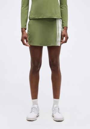 Persona che indossa una gonna verde oliva con strisce laterali bianche, maglia a maniche lunghe, calzini bianchi a coste e scarpe da ginnastica bianche, in piedi davanti a uno sfondo bianco.