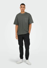 Mørkegrøn kortærmet t-shirt med brystlomme, kombineret med sorte cargo-bukser og hvide sneakers. Casual, løstsiddende design.