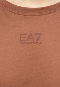 Κοντινή λήψη καφέ υφάσματος με λογότυπο EA7 Emporio Armani τυπωμένο σε αντίστοιχο καφέ χρώμα πάνω από την περιοχή του στήθους ενός πουκαμίσου.