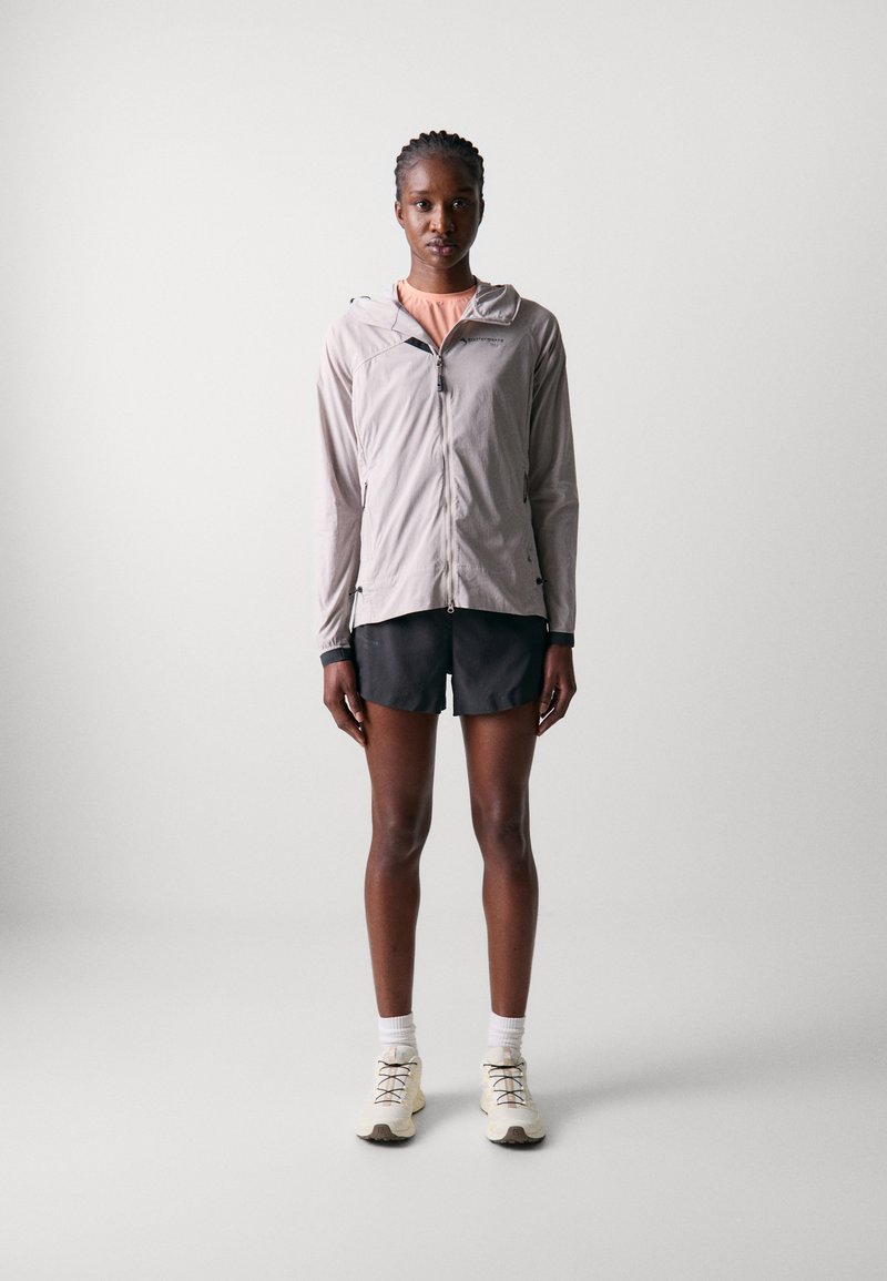 Giacca grigia leggera con chiusura a zip, colletto rialzato e cappuccio, abbinata a pantaloni corti neri e scarpe da trail beige.