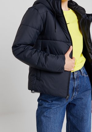 Personne portant une doudoune noire sur une chemise jaune et un jean bleu, une main ajustant la fermeture éclair de la veste.
