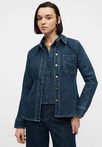 Νεαρός με κοντά μαλλιά που φοράει στρωμένη σκούρα μπλε τζιν μπουφάν και πουκάμισο πάνω από καφέ μπλουζάκι, ποζάροντας μπροστά σε απλό φόντο.