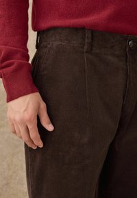 Braune CORD-Trouser mit vertikaler Ripptextur und taillierter Passform, kombiniert mit einem roten Pullover, der den Bund und die Handdetails zeigt.