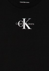Melnas kokvilnas T-krekls ar baltu "Calvin Klein Jeans" logo un stilizētu "CK" dizainu priekšā, ar apaļu kakla izgriezumu.