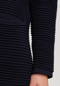 Gros plan d'une manche en maille côtelée bleu marine sur un bras, montrant des crêtes texturées horizontales et les détails du tissu.