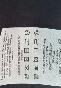 Biała metka pielęgnacyjna z czarnymi symbolami pralniczymi i tekstem w wielu językach, przymocowana do ciemnej tkaniny. Symbole wskazują instrukcje dotyczące prania, suszenia i prasowania.