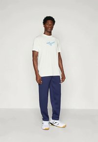 Vit t-shirt med kort ärm och blått logotyp, i kombination med marinblåa träningsbyxor och vita sneakers med gula accenter. Stående figur centrerad.