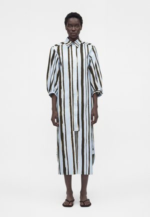 Femme debout portant une longue robe-chemise ample à rayures verticales noires et blanches et des manches bouffantes mi-longues, associée à des sandales noires.