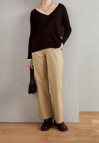 Maglione nero a scollo a V con bordi bianchi, pantaloni beige a gamba larga e stivali ankle marroni scuri. Tiene una borsa nera strutturata. Sfondo in legno.