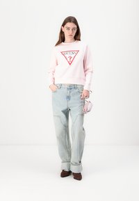 Sweatshirt rose avec logo triangulaire rouge, associé à un jean baggy bleu clair. Le modèle tient un petit sac rose ; des bottes marron complètent l'ensemble.