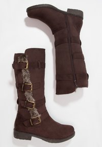Par de botas de ante marrón oscuro de altura hasta la rodilla con ribete de piel, correas con hebillas de bronce y cierre de cremallera lateral sobre un fondo blanco.