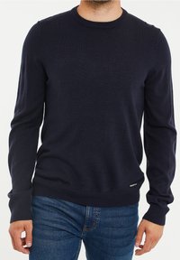 Pull en tricot bleu marine à manches longues, encolure ronde, avec poignets et ourlet côtelés. Présente un petit logo sur le côté inférieur gauche.