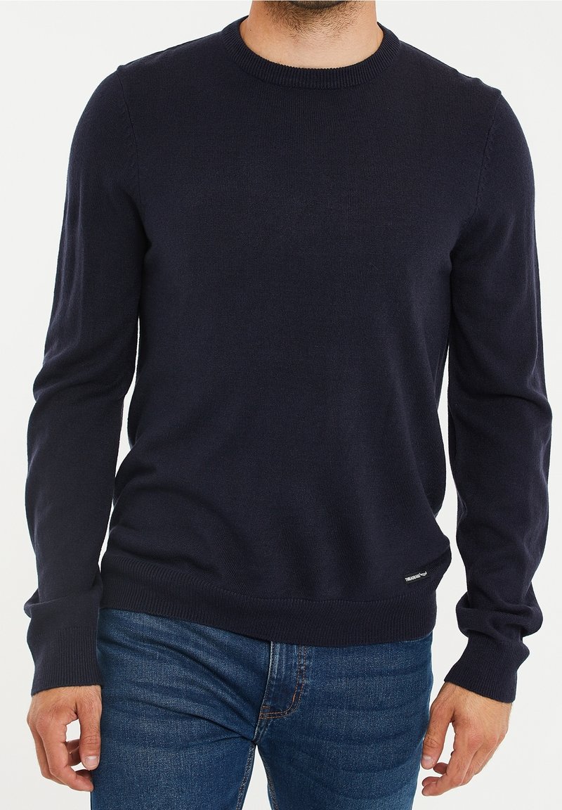 Pull en tricot bleu marine à manches longues, encolure ronde, avec poignets et ourlet côtelés. Présente un petit logo sur le côté inférieur gauche.