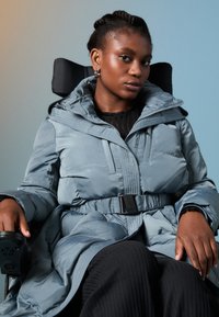 Minkštas, šviesiai mėlynas paltas su kapišonu, diržu liemeniui ir priekiniais užtrauktukais. Modelis sėdi vežimėlyje, dėvėdamas juodą, tekstūruotą marškinėlį ir kelnes.