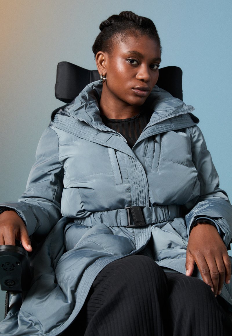 Minkštas, šviesiai mėlynas paltas su kapišonu, diržu liemeniui ir priekiniais užtrauktukais. Modelis sėdi vežimėlyje, dėvėdamas juodą, tekstūruotą marškinėlį ir kelnes.