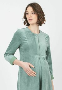 Vestido de maternidad de pana verde con escote redondo, cintura plisada y mangas de tres cuartos. Presenta un cierre de botones y tela texturizada.