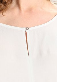 Blusa blanca con una apertura en forma de ojal en el escote, que presenta un detalle de botón plateado. Tejido ligero, suave y con una textura delicada.