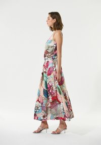 Robe maxi sans manches avec un motif floral aux couleurs vives, en tissu fluide et une taille ceinturée, accompagnée de sandales à talons blanches.