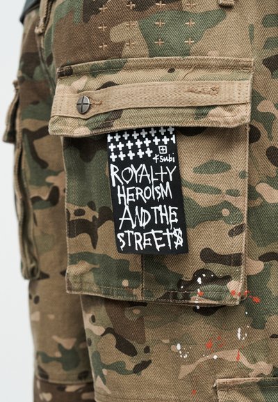 Τσέπη από παντελόνι cargo καμουφλάζ με μαύρη ετικέτα που γράφει "Βασιλεία, Ηρωισμός και οι Δρόμοι" με λευκά γράμματα σε στυλ γκράφιτι.