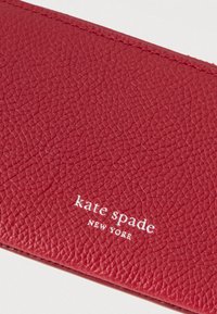 Červená textúrovaná kožená peňaženka so strieborným vyrazeným logom "kate spade NEW YORK". Hladký povrch s jemnými detailmi vzoru na povrchu.