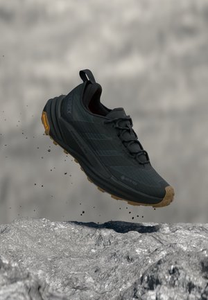 Zwarte Gore-Tex trailrunningschoen met robuuste zool, in de lucht boven rotsachtig terrein, vuildeeltjes zwevend rond de schoen.