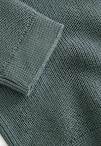 Grøn ribbet strikket stof med en tekstureret overflade, der har en folded cuff-detalje og et ensartet horisontalt mønster gennem hele stoffet.
