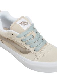 Béžové tenisky Vans se světle modrými tkaničkami, bílou podrážkou a logem "Vans Off The Wall" na jazyku, zobrazené z mírného úhlu.