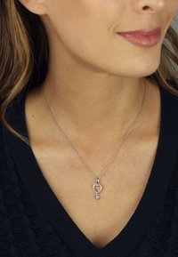 Silberne Kettenhalskette mit einem herzförmigen Anhänger, an dem ein kleiner dekorativer Stein hängt. Texturiertes dunkles Oberteil darunter getragen.