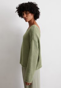 Übergroßer grüner Strickpullover mit strukturiertem Muster, weitem Ausschnitt und langen, lockeren Ärmeln. Kombiniert mit hellen Hosen.