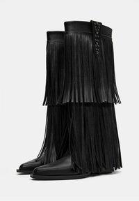 Črne usnjene škornje do kolen z dolgimi resicami vzdolž zgornjega dela in študiranimi detajli na vrhu za dodaten tekstur.