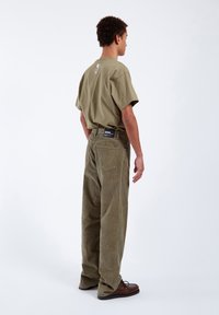 Ung mand står med ryggen til iført en olivengrøn T-shirt med hjerte-designs og matchende kordfløjlsbukser med brune sko.