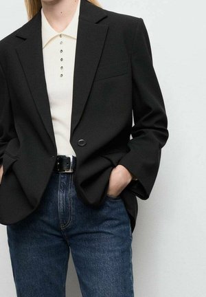 Person iført sort blazer over hvid skjorte med knapper, stoppet ned i blå jeans med sort bælte, hænder i lommerne, mod en ensartet lys baggrund.