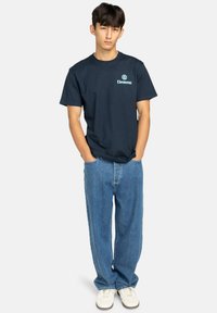Marineblaues Baumwoll-T-Shirt mit einem hellblauen "Element"-Logo auf der Brust, kombiniert mit lässig sitzenden Jeans und weißen Turnschuhen.