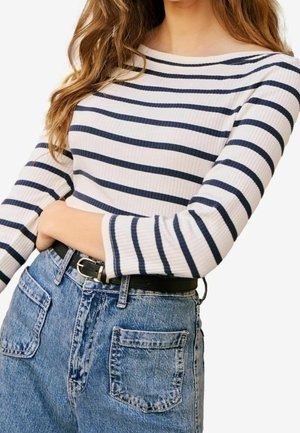 Femme portant un haut à manches longues blanc et bleu marine à rayures, rentré dans un jean bleu taille haute avec une ceinture noire.