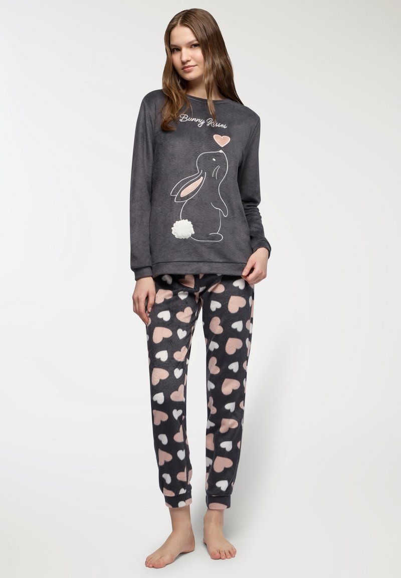 Pigiama di pile grigio con un grafico di coniglio e una coda bianca fluffosa; pantaloni decorati con motivi a cuori rosa e bianchi.