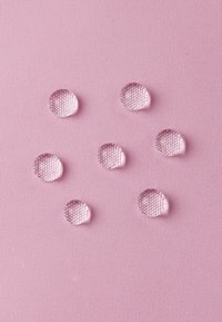 Gocce d'acqua trasparenti su una superficie rosa chiaro e testurizzata, che mostrano una finitura liscia e lucida e una forma circolare. Sei gocce visibili.