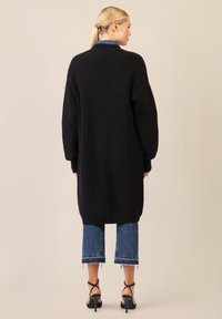 Cardigan lungo nero in maglione con texture a coste, spalle scese e vestibilità over. Abbinato a jeans in denim a vita alta e sandali neri con cinturini.