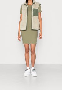 Abito bodycon khaki abbinato a un gilet morbido crema con tasca verde e sneakers bianche con accenti grafici multicolori.