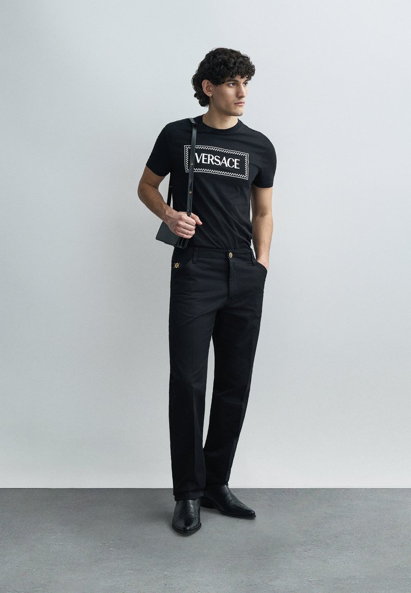 Svart t-shirt med "VERSACE"-logotyp, kombinerad med svarta byxor med en gulddetalj. Modellen håller en liten axelväska och bär svarta skor.