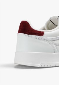 White sneaker with a textured leather upper, red suede heel accent, and distinct white rubber sole with horizontal grooves.