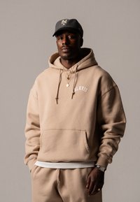 Hanorac din bumbac bej cu buzunar frontal, șnur și logo "BLKVIS". Asortat cu pantaloni de trening bej. Capac gri cu accent de logo.