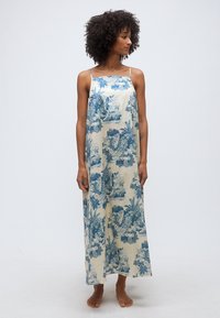 Γυναίκα με σγουρά μαλλιά, φοράει αμάνικο κρεμ μακρύ φόρεμα με μπλε λουλουδάτα και τοπία τύπωμα, όρθια ξυπόλητη σε απλό φόντο.