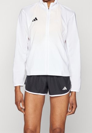 Osoba nosząca białą sportową kurtkę na zamek błyskawiczny oraz czarne spodenki do biegania Adidas z białymi lamówkami na jednolitym tle.